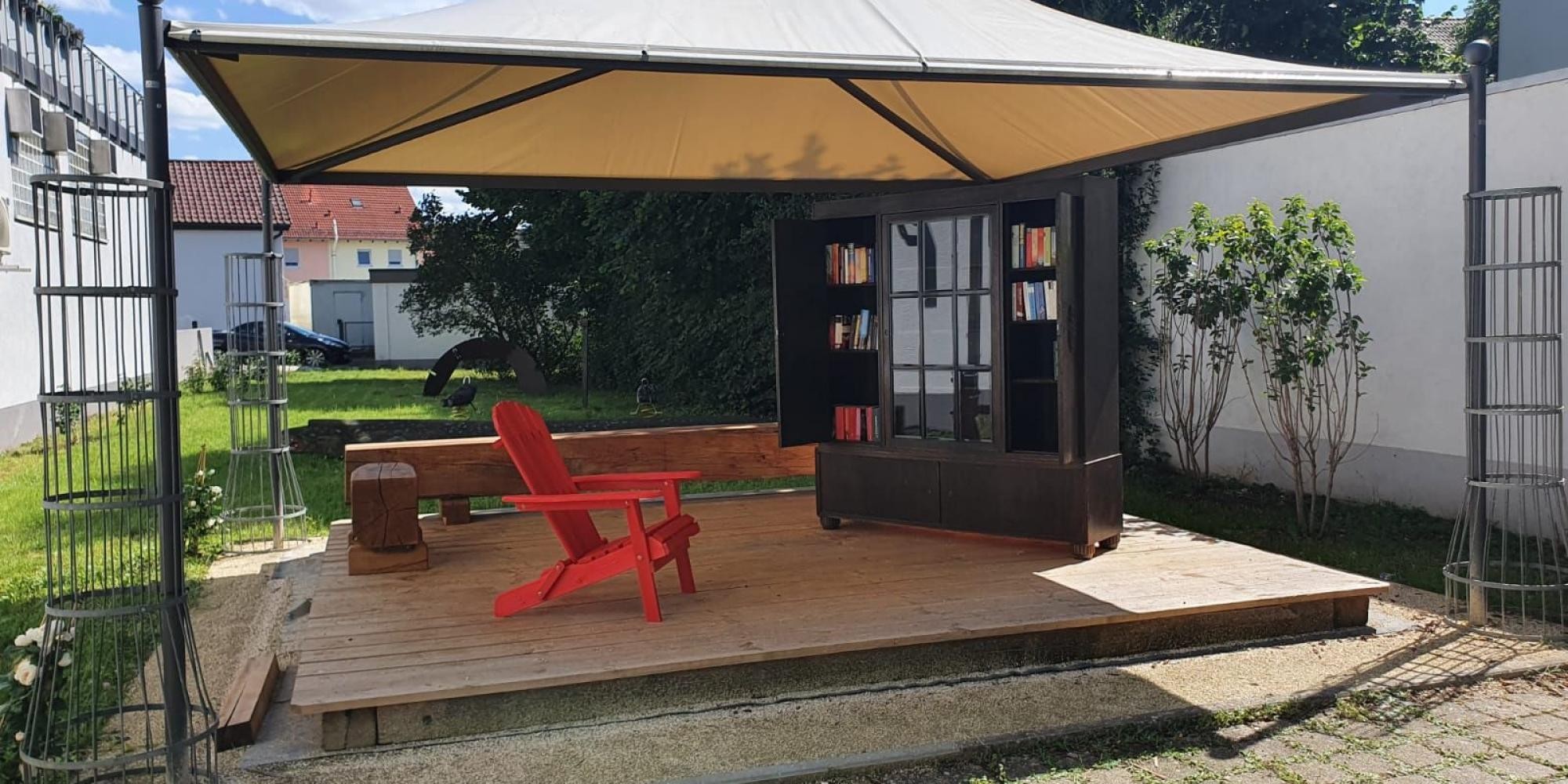 Großer Pavillon, darunter ein roter Stuhl, dahinter Sitzbänke, rechts ein Bücherregal