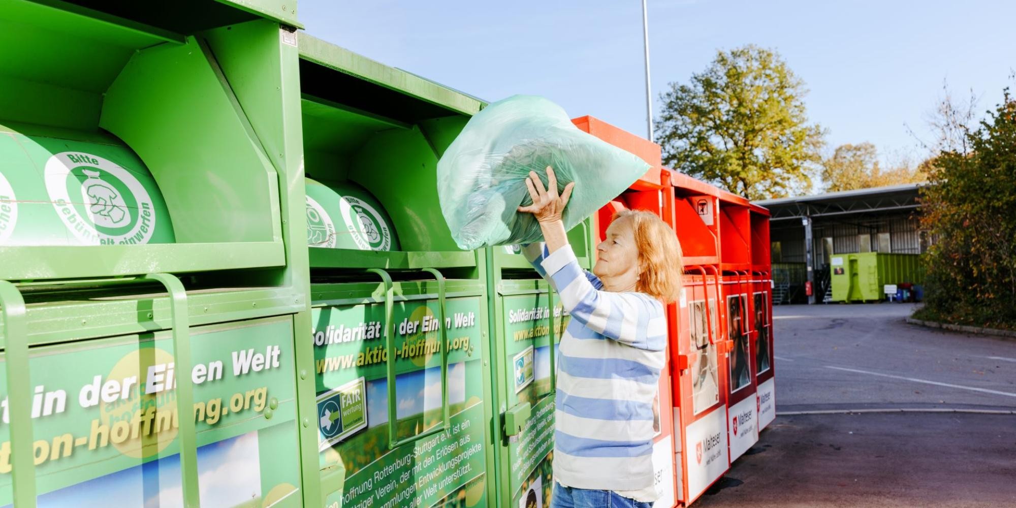 Mehrere Altkleidercontainer stehen nebeneinander. Eine Frau wirft einen Sack mit Alttextilien in einen der Container.