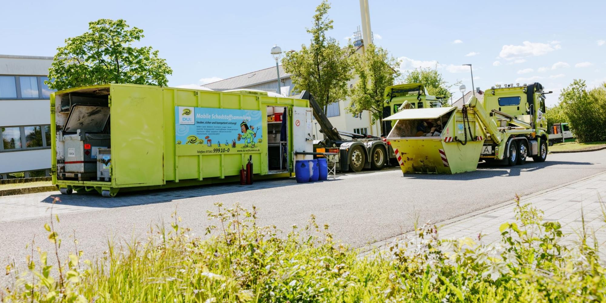 Auf einem großen Parkplatz stehen zwei LKW, hinter denen jeweils ein Container abgestellt, aber noch am Greifarm eingehängt ist. Der linke Container ist begehbar, davor ist ein Tisch aufgebaut und drei blaue Kunststofffässer stehen bereit. Beim rechten Container handelt es sich um eine Mulde mit Deckel, der leicht geöffnet ist.
