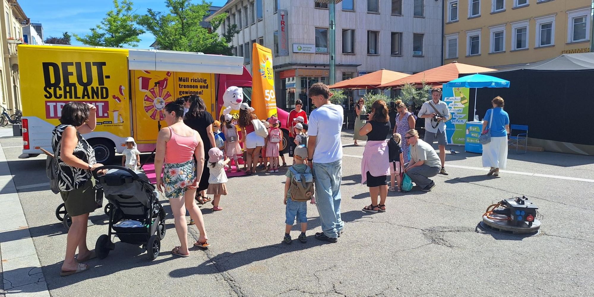 Vor dem Showtruck der Aktion "Deutschland trennt" steht der Trennbär und das Fragenrad. Davor stehen einige Kinder und Erwachsene. Am Rand befindet sich der Infostand des Abfallwirtschaftsbetriebs.