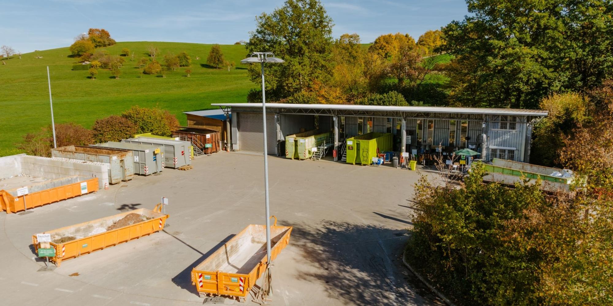 Drohnenaufnahme des Wertstoffzentrums, im Vordergrund drei Bauschuttmulden, im hinteren Teil überdachter Bereich mit weiteren Containern, im Hintergrund Wiese und Bäume.
