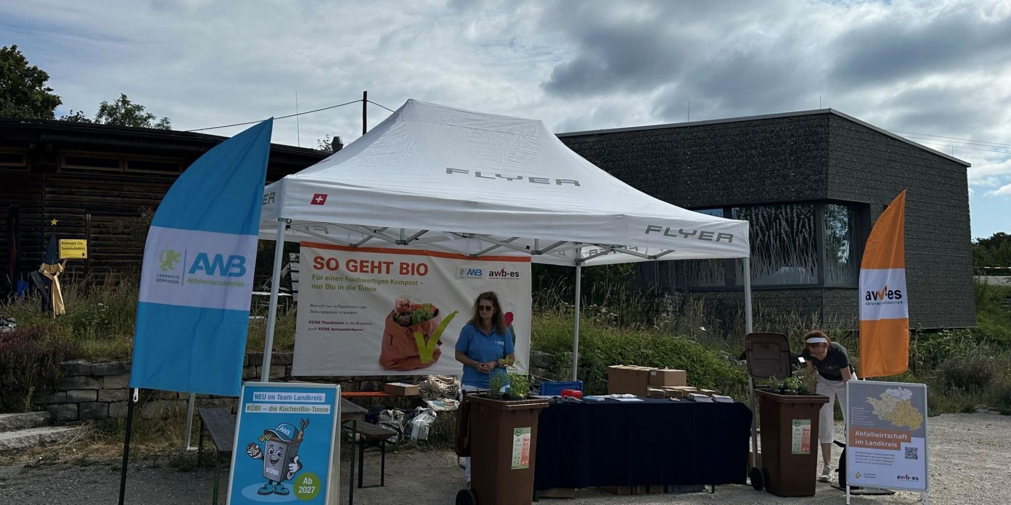 Mitarbeiterin an einem Infostand unter einem großen Pavillon. Rechts und links stehen Beachflags und Informationsaufsteller. Zwei bepflanzte Mülltonnen rahmen den Stand ein. Im Hintergrund hängt ein großes Banner mit der Aufschrift „So geht Bio“.