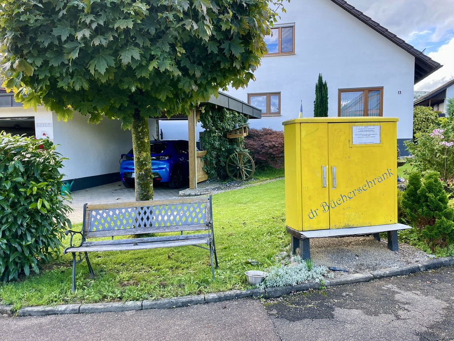 Ein gelb gestrichener Schaltschrank mit der Aufschrift "dr Bücherschrank", links daneben eine Sitzbank vor einem Baum.