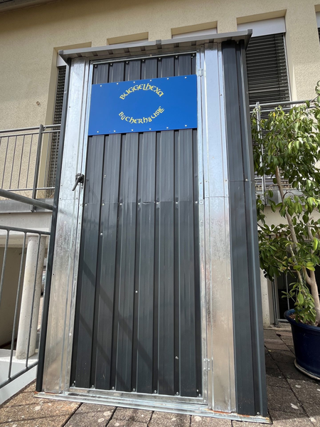 Bücherschrank, auf dem ein blaues Schild mit der Aufschrift "Buggelhexa Bücherhäusle" angebracht ist.