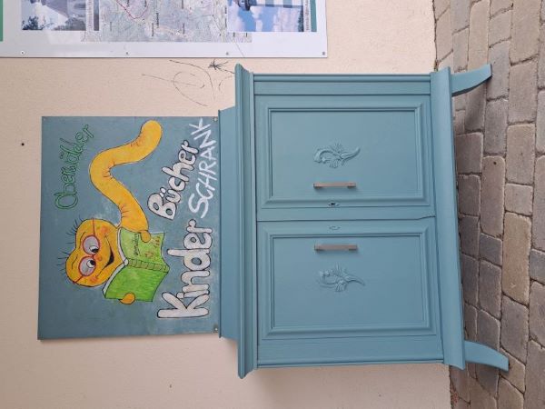 Blaue Kommode. Darüber hängt ein blaues Brett, auf das ein gelber Bücherwurm aufgemalt ist, darunter steht "Kinderbücherschrank".